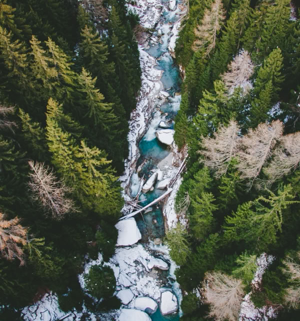 Aerial view of a narrow river flowing through a forest with snow-covered rocks and trees.